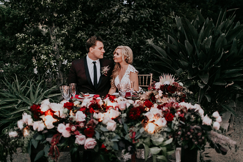 orange-county-vow-renewal-bride-and-groom-at-sweetheart-table-bride-in-a-lace-modern-fitted-gown-with-a-nude-underlay-groom-in-a-burgundy-tuxedo-with-a-black-shawl-lapel