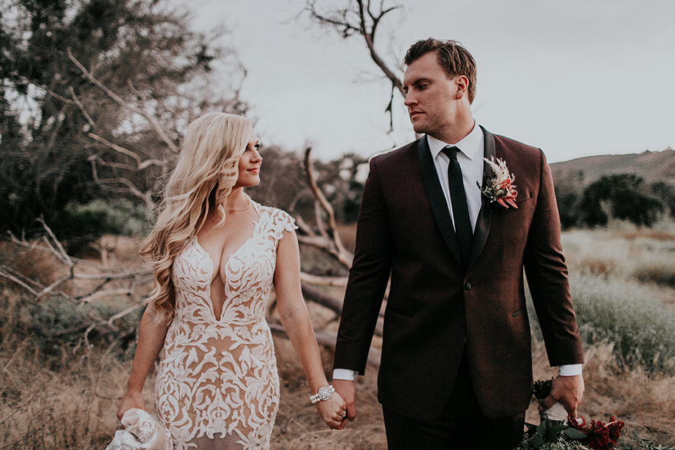 orange-county-vow-renewal-brode-and-groom-walking-and-looking-at-each-other-bride-in-a-lace-modern-fitted-gown-with-a-nude-underlay-groom-in-a-burgundy-tuxedo-with-a-black-shawl-lapel