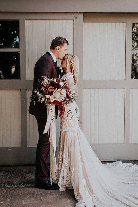 orange-county-vow-renewal-groom-kissing-bride-on-forehead-bride-in-a-lace-modern-fitted-gown-with-a-nude-underlay-groom-in-a-burgundy-tuxedo-with-a-black-shawl-lapel