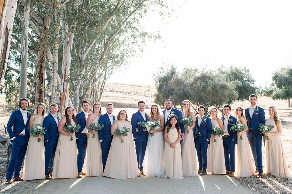 bride in a white a line gown with straps and a big white and sage floral bouquet and cathedral length veil, the groom in a dark blue suit with a white bow tie, the groomsmen in blue suits and champagne ties and the bridesmaids in neutral long gowns