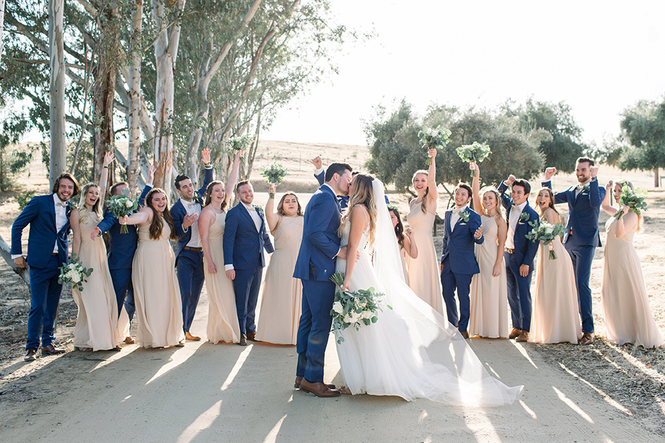  bride in a white a line gown with straps and a big white and sage floral bouquet and cathedral length veil, the groom in a dark blue suit with a white bow tie, the groomsmen in blue suits and champagne ties and the bridesmaids in neutral long gowns