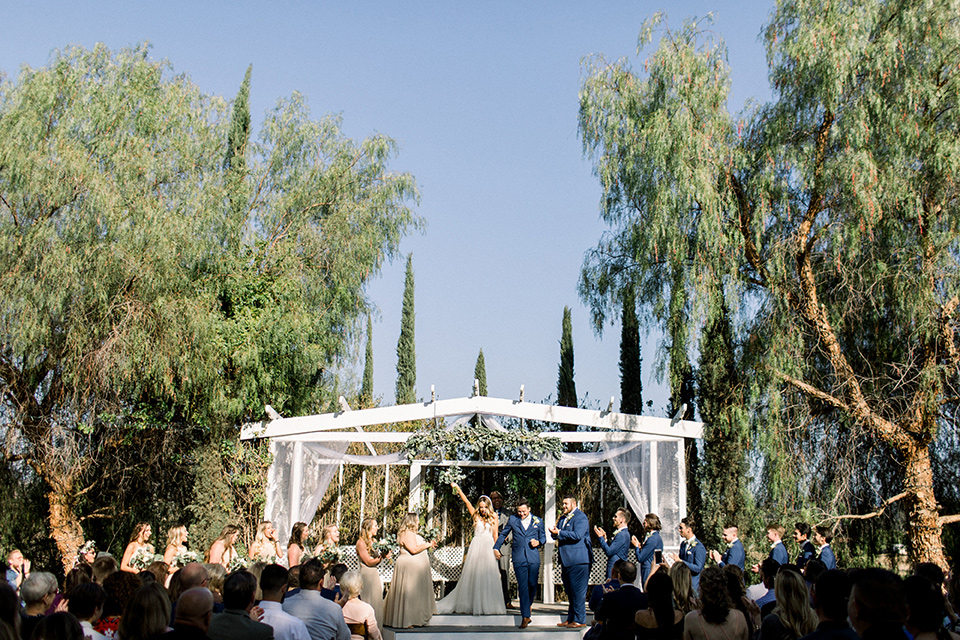  bride in a white a line gown with straps and a big white and sage floral bouquet and cathedral length veil, the groom in a dark blue suit with a white bow tie, the groomsmen in blue suits and champagne ties and the bridesmaids in neutral long gowns all under a white awning and surrounded by trees