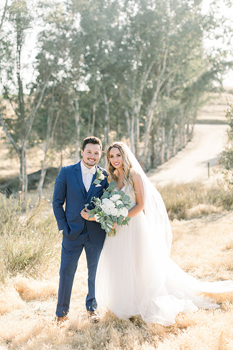  bride in a white a line gown with straps and a big white and sage floral bouquet and cathedral length veil, the groom in a dark blue suit with a white bow tie