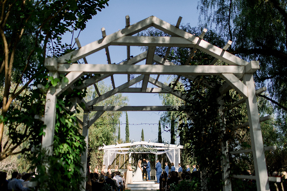  bride in a white a line gown with straps and a big white and sage floral bouquet and cathedral length veil, the groom in a dark blue suit with a white bow tie, the groomsmen in blue suits and champagne ties and the bridesmaids in neutral long gowns all under a white awning and surrounded by trees