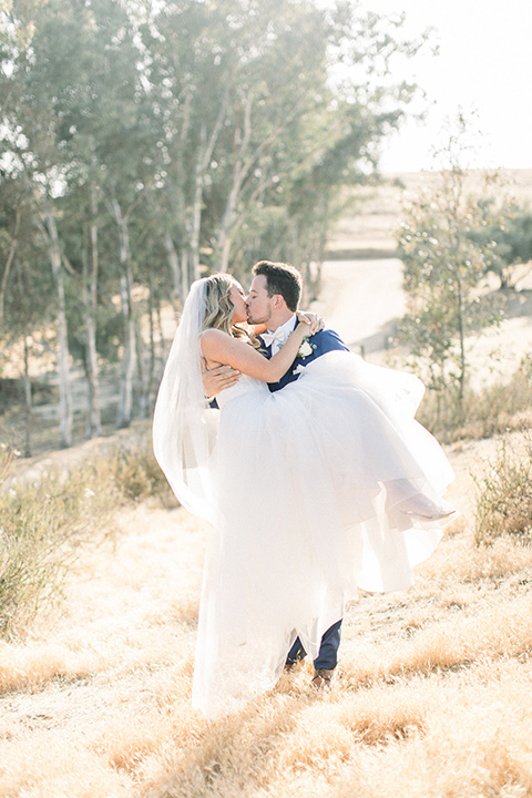  bride in a white a line gown with straps and a big white and sage floral bouquet and cathedral length veil, the groom in a dark blue suit with a white bow tie 