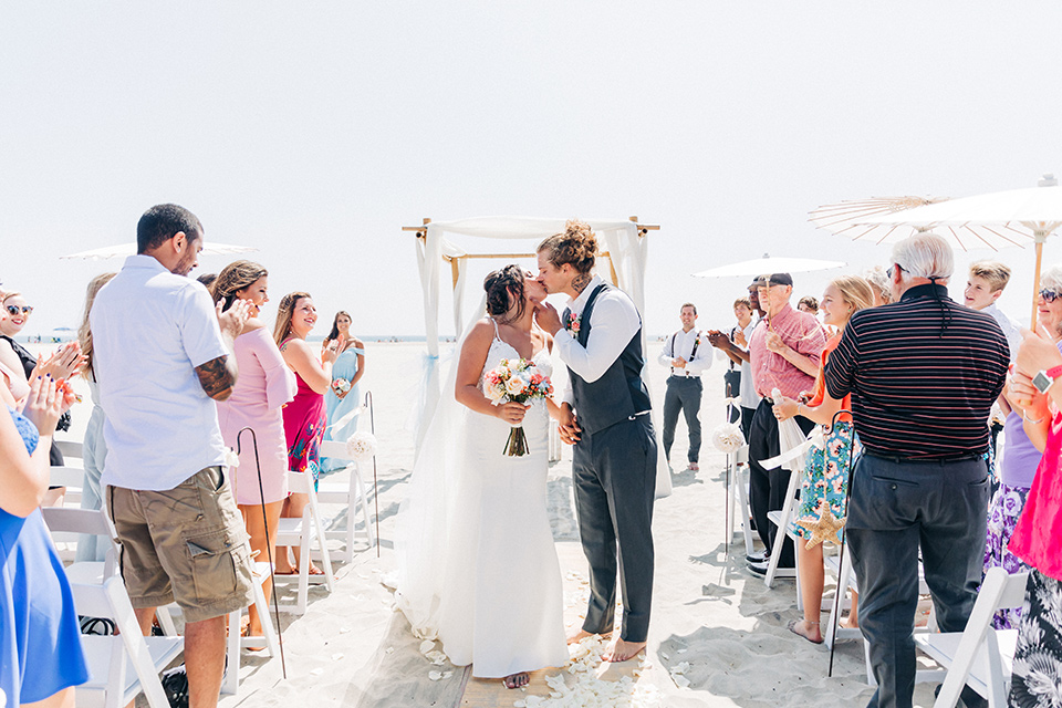 San-Diego-Beach-wedding-bride-and-groom-kissing-at-end-of-aisle-bridesmaids-in-bright-blue-dresses-groomsmen-in-suit-pants-and-suspenders-bride-ina-lace-formitting-dress-with-strapsand-a-veil-groom-in-a-suit-pants-and-vest