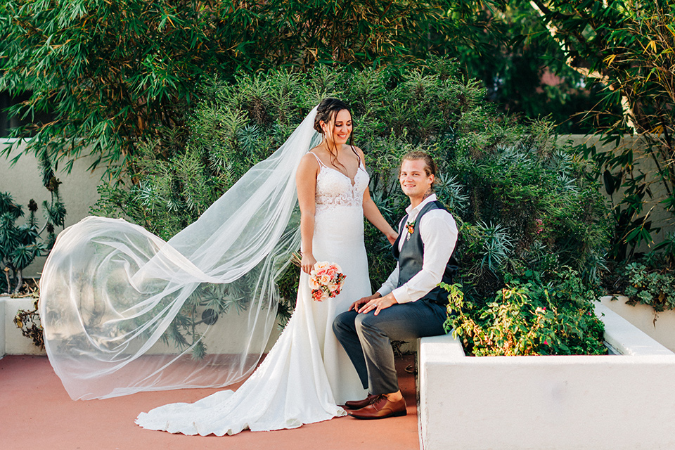 San-Diego-Beach-wedding-groom-sitting-brode-stands-with-veil-in-the-wind-bride-ina-lace-formitting-dress-with-straps-and-a-veil-groom-in-a-suit-pants-and-vest