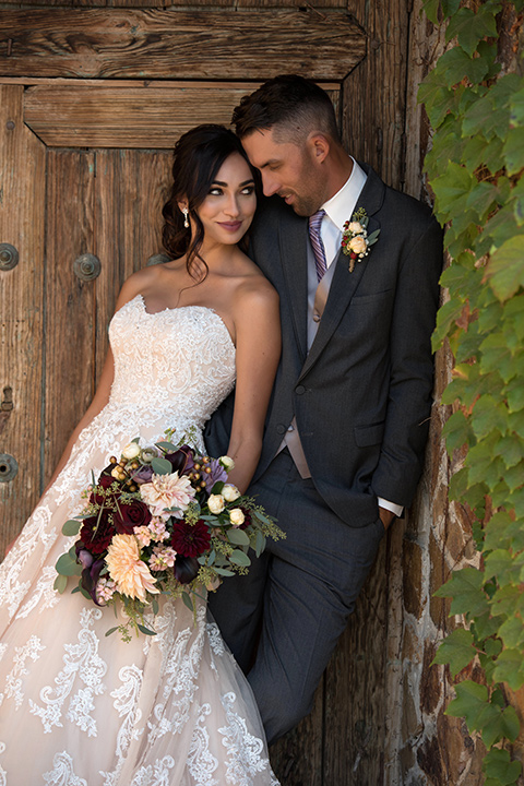 milagro-farms-shoot-bride-and-groom-leaning-against-each-other-by-door-bride-in-a-fitted-lace-gown-with-straps-groom-in-a-charcoal-suit-with-a-purple-tie