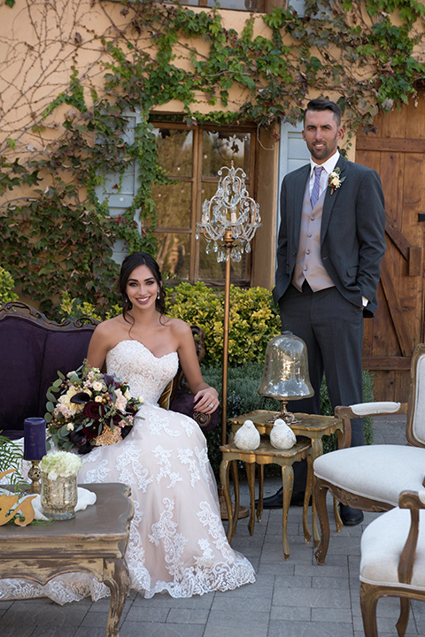 milagro-farms-shoot-bride-sitting-groom-standing-bride-in-a-fitted-lace-gown-with-straps-groom-in-a-charcoal-suit-with-a-purple-tie