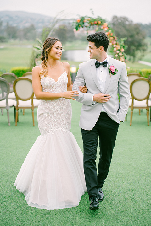 omni-la-costa-bride-and-groom-walking-down-ceremony-bride-wearing-a-lace-fitted-gown-with-mermaid-style-with-thin-straps-groom-with-light-grey-jacket-with-black-pants-black-polka-dot-bowtie