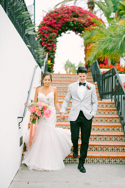 omni-la-costa-bride-and-groom-walking-down-the-stairs-bride-wearing-a-lace-fitted-gown-with-mermaid-style-with-thin-straps-groom-with-light-grey-jacket-with-black-pants-black-polka-dot-bowtie