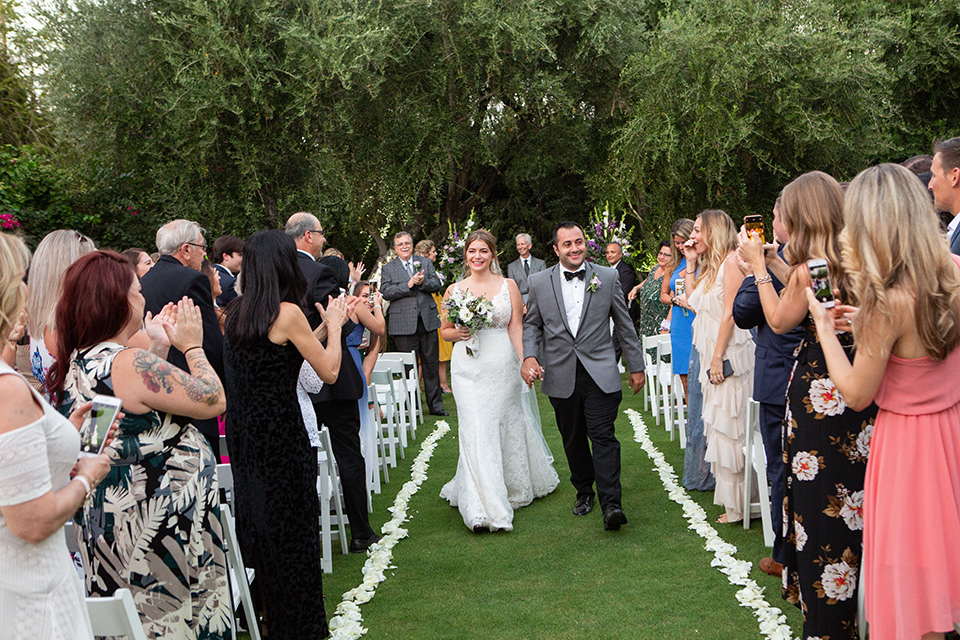 Palm-Springs-wedding-at-the-parker-bride-and-groom-walking-down-the-aisle-bride-in-a-flowing-gown-with-straps-and-a-plunging-deep-v-neckline-groom-in-a-grey-tuxedo-with-black-trim-and-black-pants