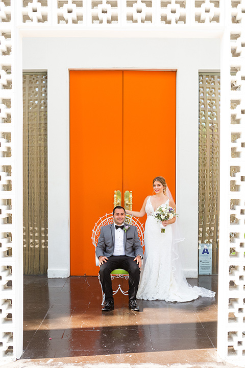 Palm-Springs-wedding-at-the-parker-groom-sitting-bride-standing-bride-in-a-flowing-gown-and-a-deep-v-neckline-groom-in-a-grey-tux-with-black-trim-and-black-pants