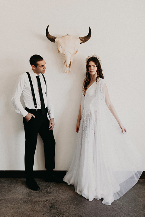 Rim-Rock-Ranch-Shoot-bride-and-groom-by-longhorn-skull-bride-wearing-a-sheer-gown-with-crystals-in-it-with-flutter-sleeves-and-a-crown-groom-in-a-white-shirt-black-pants-and-black-suspenders