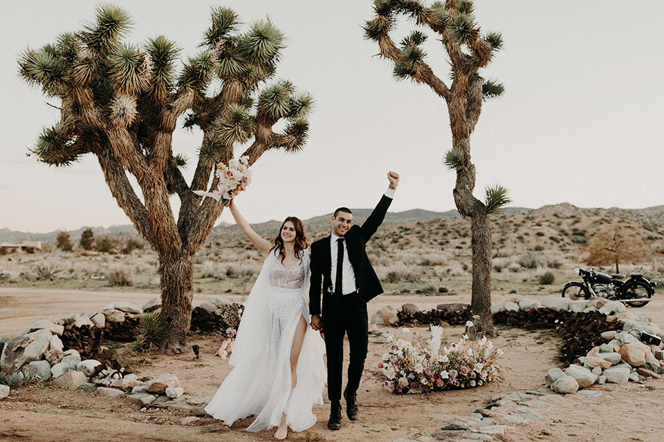 Rim-Rock-Ranch-Shoot-bride-and-groom-in-ceremony-space-with-hands-in-the-air-bride-in-a-bohemian-style-dress-with-a-sheer-overlay-and-crown-groom-in-a-simple-black-suit-and-long-tie