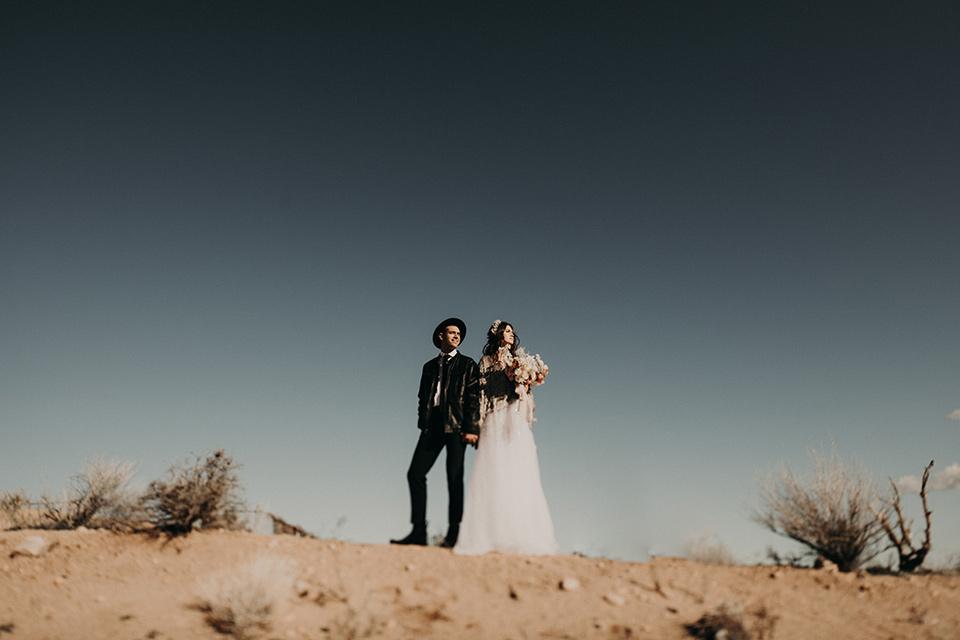 Rim-Rock-Ranch-Shoot-bride-and-groom-on-hilltop-with-hat-on-bride-in-a-bohemian-style-dress-with-a-sheer-overlay-and-crown-groom-in-a-simple-black-suit-and-long-tie