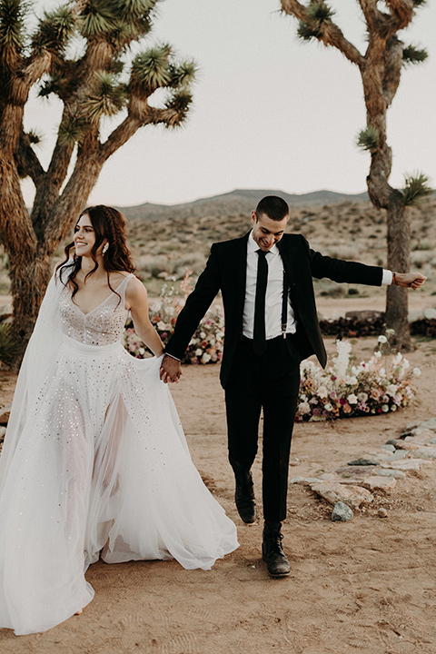 Rim-Rock-Ranch-Shoot-bride-and-groom-walking-bride-wearing-a-sheer-gown-with-crystals-in-it-with-flutter-sleeves-and-a-crown-groom-in-a-white-shirt-black-pants-and-black-suspenders