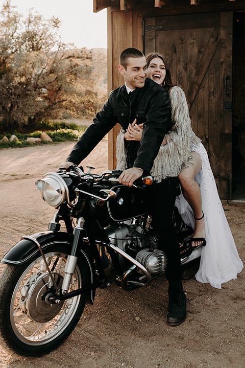 Rim-Rock-Ranch-Shoot-bride-laughing-on-motocycle-groom-steering-bride-wearing-a-sheer-gown-with-crystals-in-it-with-flutter-sleeves-and-a-crown-groom-in-a-white-shirt-black-pants-and-black-suspenders