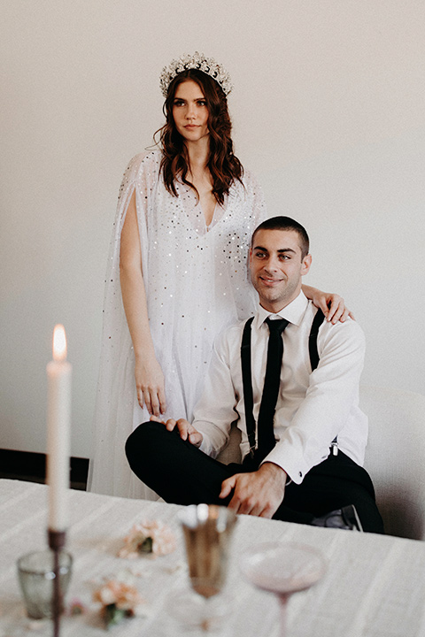 Rim-Rock-Ranch-Shoot-groom-sitting-bride-standing-bride-with-crown-on-and-a-sheer-bohemian-dress-with-crsystals-and-flutter-sleeves-groom-in-a-white-shirt-black-pants-and-black-suspenders