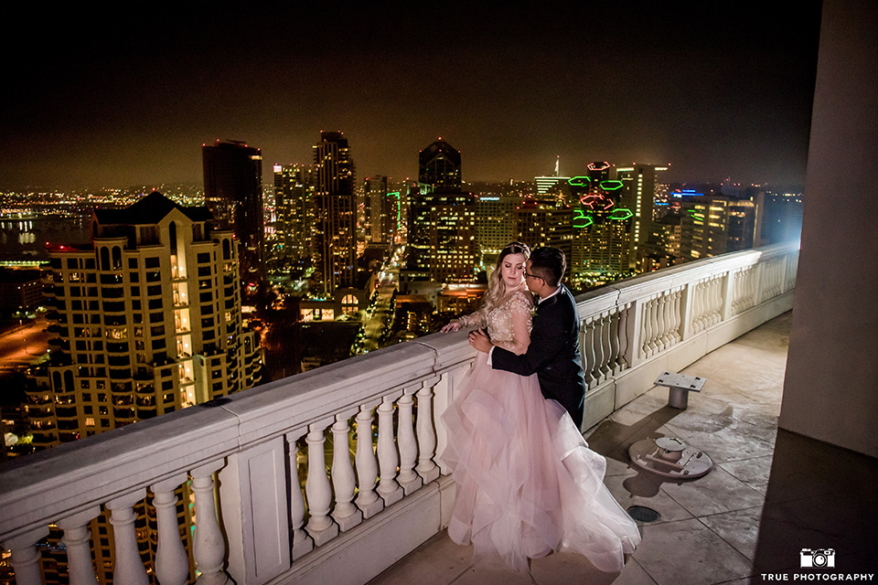 Hyatt-San-Diego-wedding-bride-and-groom-overlooking-city-at-night-the-bride-is-in-a-blush-toned-ballgown-and-the-groom-in-a-black-shawl-lapel-tuxedo