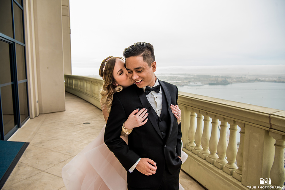 Hyatt-San-Diego-wedding-bride-hugging-groom-from-behind-the-bride-is-in-a-blush-toned-ballgown-and-the-groom-in-a-black-shawl-lapel-tuxedo