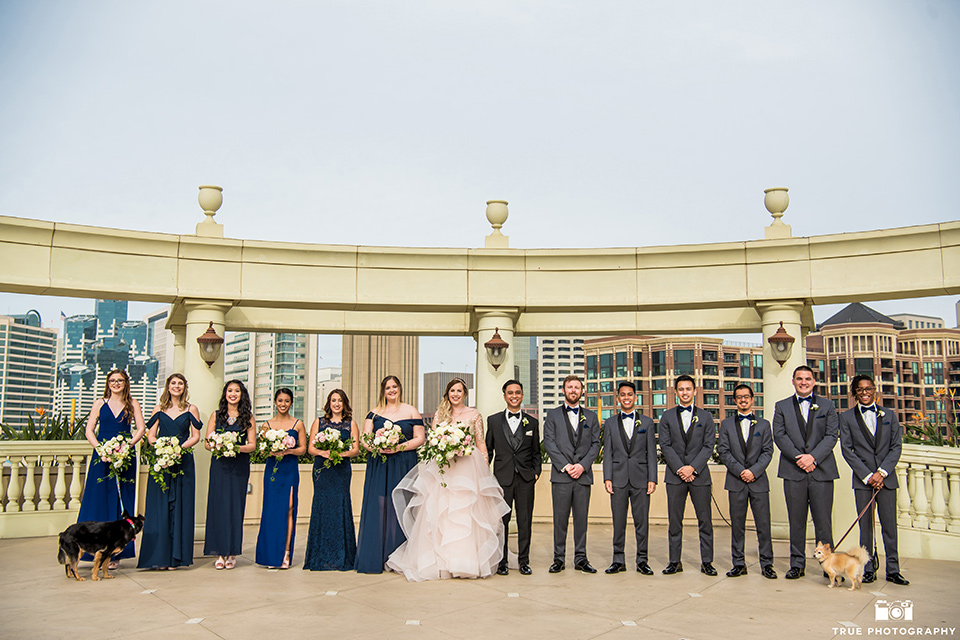 Hyatt-San-Diego-wedding-bridalparty-the-bridesmaids-in-navy-blue-gowns-groomsmen-in-grey-tuxedos-the-bride-is-in-a-blush-toned-ballgown-and-the-groom-in-a-black-shawl-lapel-tuxedo