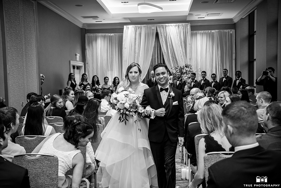 hyatt-san-diego-wedding-bride-and-groom-walking-down-aisle-the-bride-is-in-a-blush-toned-ballgown-and-the-groom-in-a-black-shawl-lapel-tuxedo