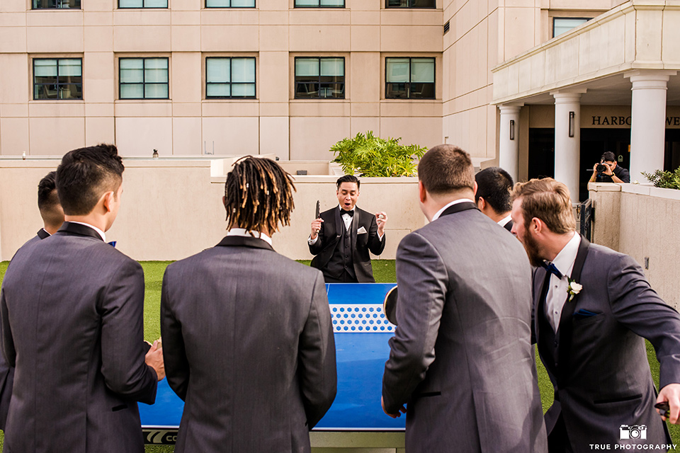 hyatt-san-diego-wedding-groomsmen-looking-at-grrom-playing-ping-pong-the-groom-in-a-black-shawl-lapel-tuxedo-the-groomsmen-in-grey-shalw-lapel-tuxedos