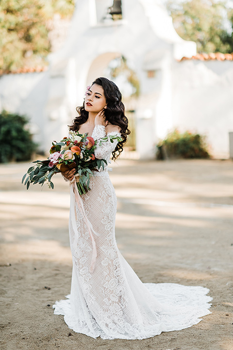 Olivas-Adobe-spanish-inspired-shoot-bride-alone-touching-her-neck-wearing-a-lace-form-fitting-gown-with-off-the-shoulder-detailing-and-long-sleeves