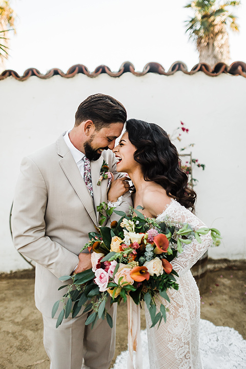 Olivas-Adobe-spanish-inspired-shoot-bride-and-groom-laughing-bride-wearing-a-lace-form-fitting-gown-with-off-the-shoulder-detailing-and-long-sleeves-groom-wearing-a-tan-suit-with-a-white-floral-neck-tie