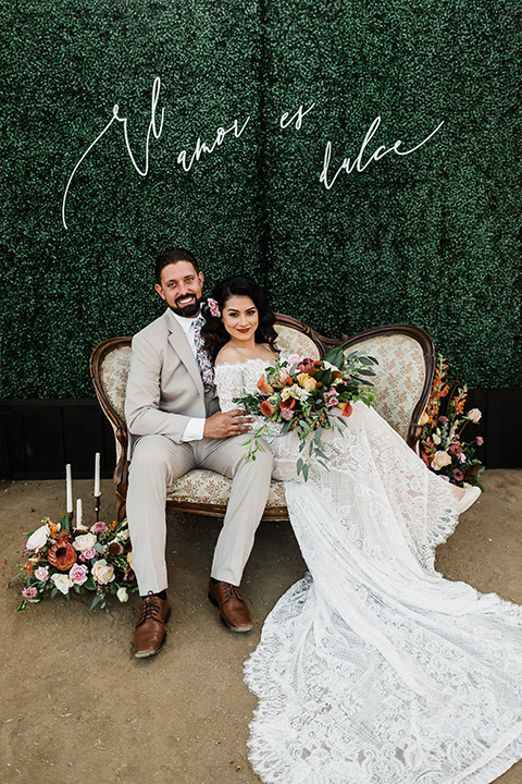 Olivas-Adobe-spanish-inspired-shoot-bride-and-groom-sitting-bride-wearing-a-lace-form-fitting-gown-with-off-the-shoulder-detailing-and-long-sleeves-groom-wearing-a-tan-suit-with-a-white-floral-neck-tie