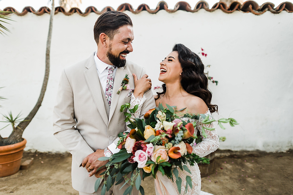 Olivas-Adobe-spanish-inspired-shoot-bride-and-groom-smiling-bride-in-a-spanish-inspired-lace-dress-with-long-sleeves-and-groom-in-a-tan-suit-with-a-floral-tie