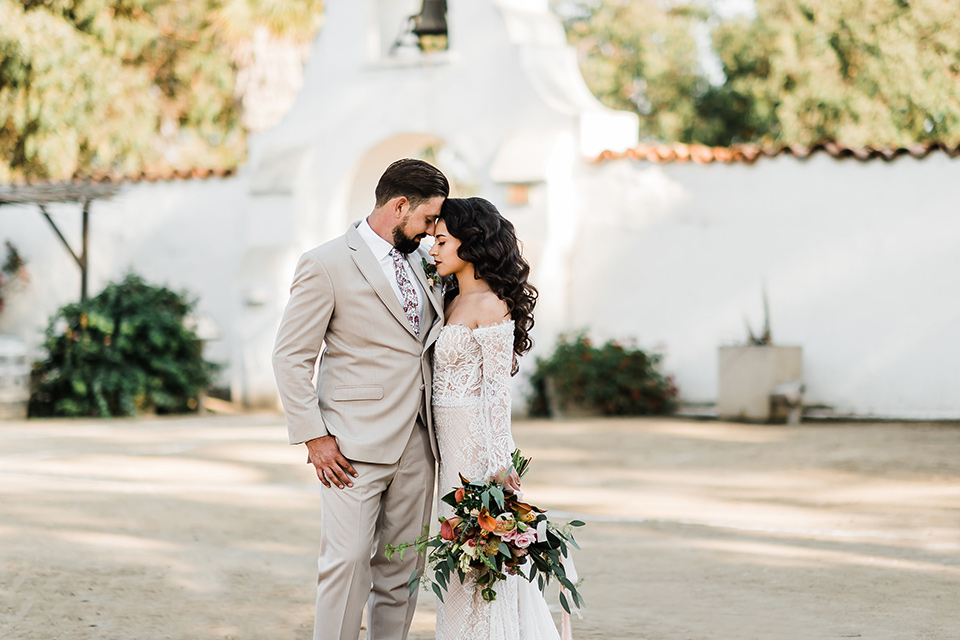 Olivas-Adobe-spanish-inspired-shoot-bride-and-groom-touching-heads-bride-in-a-spanish-inspired-lace-dress-with-long-sleeves-and-groom-in-a-tan-suit-with-a-floral-tie