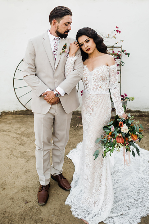 Olivas-Adobe-spanish-inspired-shoot-couple-looking-serious-bride-wearing-a-lace-gown-with-sleeves-and-holding-a-large-bouquet-of-flowers-groom-wearing-a-tan-suit-with-a-floral-tie