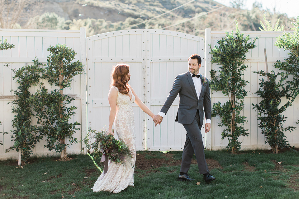 triunfo-creek-vineyards-wedding-bride-and-groom-walking-by-fence-bride-in-a-lace-gown-in-a-blush-tone-groom-in-a-grey-tuxedo-with-a-black-bow-tie