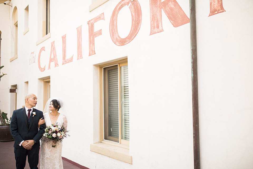 villa-del-sol-wedding-couple-walking-by-venue-bride-in-a-spanish-inspired-lace-gown-with-a-deep-v-and-long-sleeves-groom-in-a-navy-shawl-lapel-tuxedo-with-a-burgundy-tie