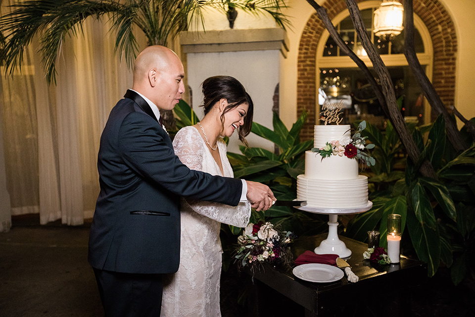 villa-del-sol-wedding-cutting-the-cake-bride-in-a-spanish-inspired-lace-gown-with-a-deep-v-and-long-sleeves-groom-in-a-navy-shawl-lapel-tuxedo-with-a-burgundy-tie