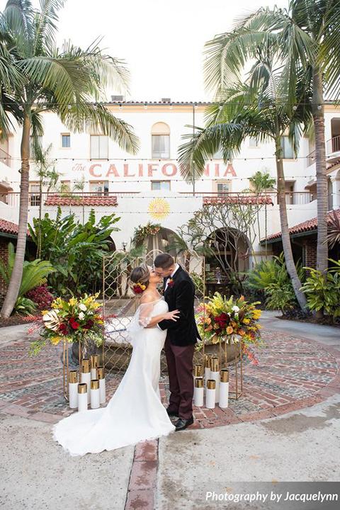  bride wearing white pointed toe shoes and a white long gown with flowing sleeves and a high neckline and the groom in a black velvet tuxedo by fountain at venue