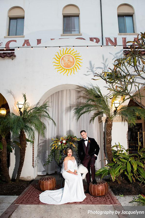  bride in a white silk gown with flowing sleeves and a high neckline and the groom in a black velvet tuxedo sitting in venue