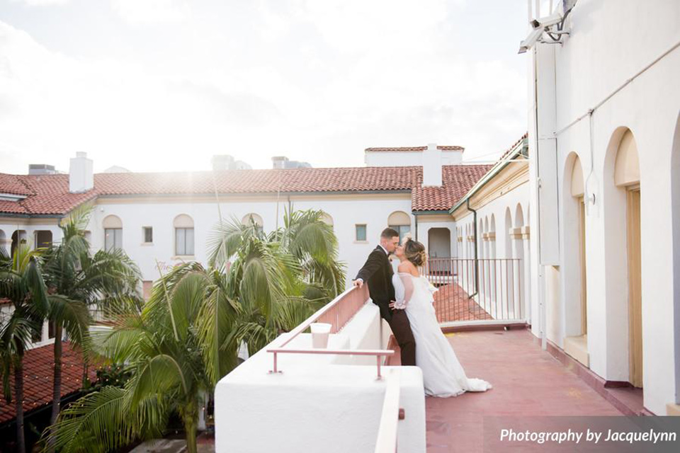  bride in a white silk gown with flowing sleeves and a high neckline and the groom in a black velvet tuxedo on balcony