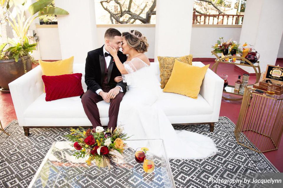  bride in a white silk gown with flowing sleeves and a high neckline and the groom in a black velvet tuxedo sitting on couch