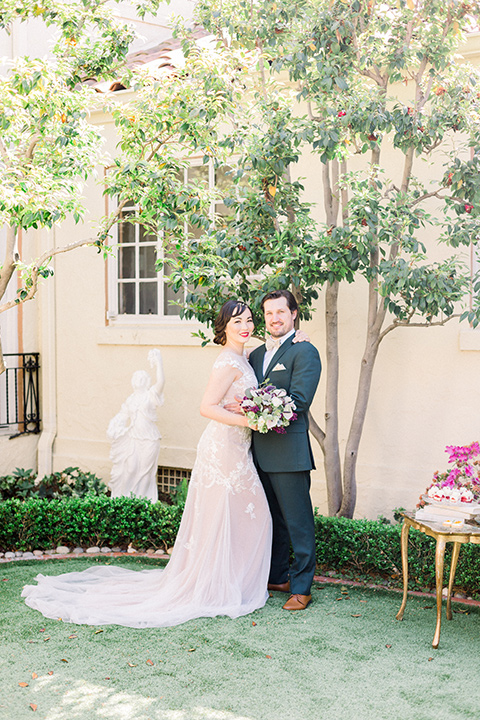  bride in a Spanish style white lace gown with sleeves and deep red lipstick and the groom in a dark green suit with a tan bow tie and brown shoes