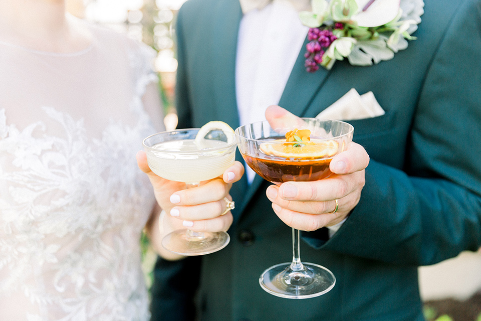  bride in a Spanish style white lace gown with sleeves and dee red lipstick and the groom in a dark green suit with a tan bow tie and brown shoes holding cocktails