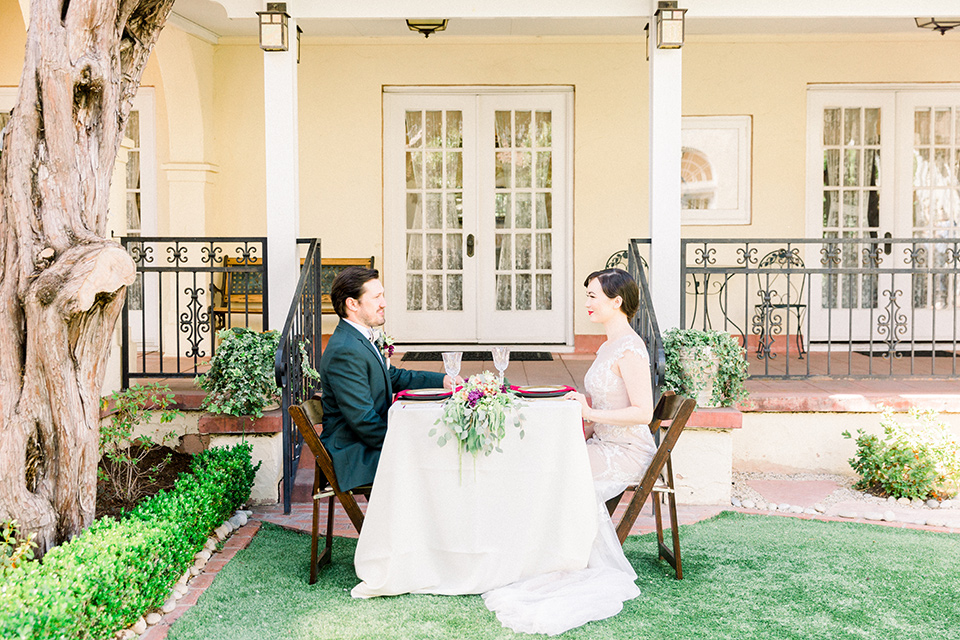  bride in a Spanish style white lace gown with sleeves and dee red lipstick and the groom in a dark green suit with a tan bow tie and brown shoes holding flowers