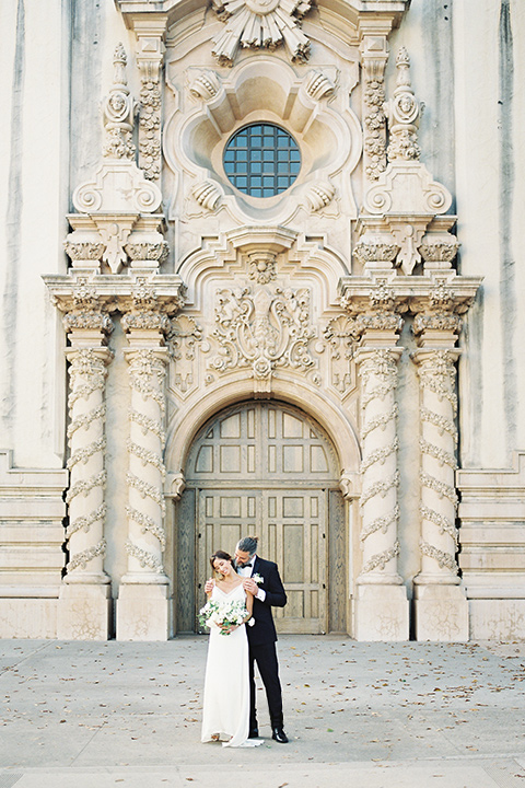  bride in a satin formfitting gown with thin straps, and the groom in a black notch lapel tuxedo with a black bow tie 