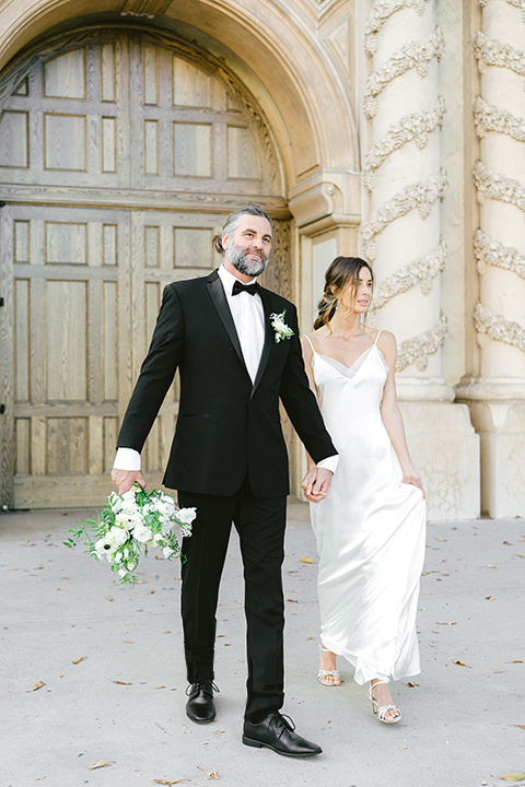  bride in a satin formfitting gown with thin straps, and the groom in a black notch lapel tuxedo with a black bow tie