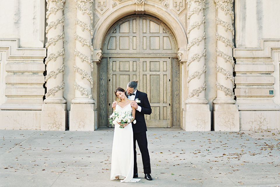  bride in a satin formfitting gown with thin straps, and the groom in a black notch lapel tuxedo with a black bow tie 