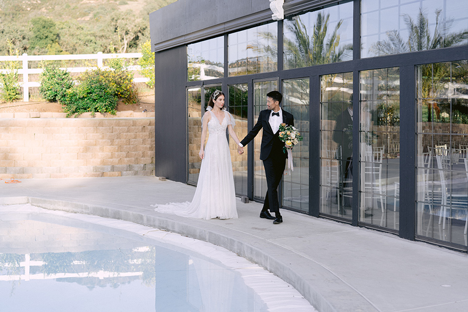  bride in a white lace gown with a flowing skirt with a high slit on the side, and tie straps.  The groom in a black michael kors tuxedo with a black bow tie 