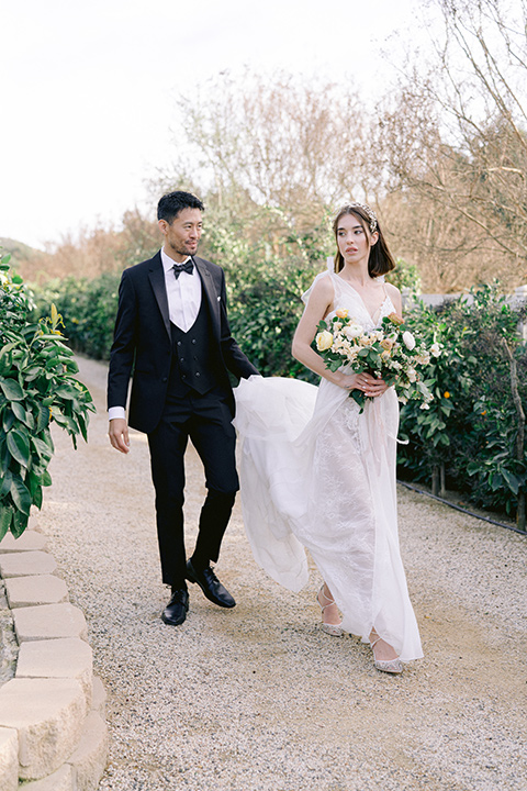  bride in a white lace gown with a flowing skirt with a high slit on the side, and tie straps.  The groom in a black michael kors tuxedo with a black bow tie 