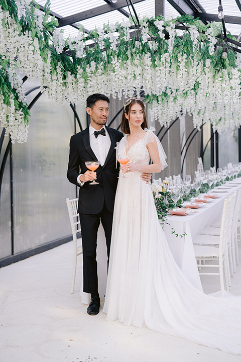  bride in a white lace gown with a flowing skirt with a high slit on the side, and tie straps.  The groom in a black michael kors tuxedo with a black bow tie 